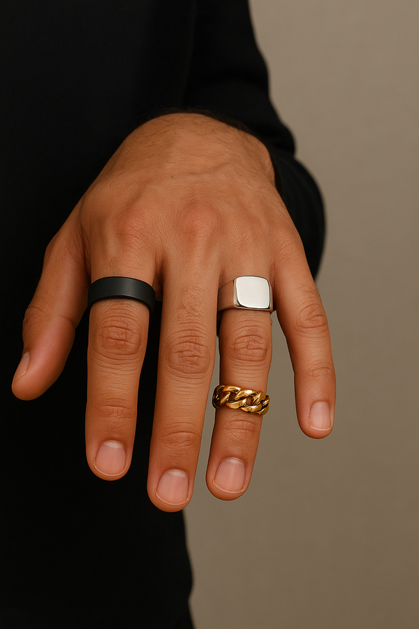 Close-up of a man's hand wearing three stylish rings: a matte black band, a silver signet, and a gold Cuban ring.