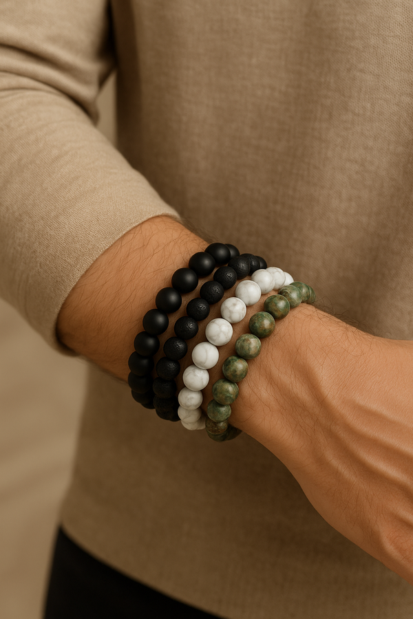 A close-up of a man's wrist wearing five stylish bracelets — a gold cuff, matte black agate, white howlite, lava stone, and green jasper — styled with a beige shirt, showcasing trendy men's wristwear for 2025.