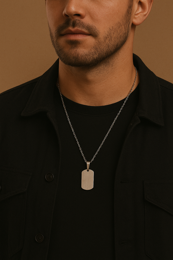 A close-up of a man wearing a black t-shirt and jacket with a silver dog-tag style pendant hanging from a chain against a beige background.