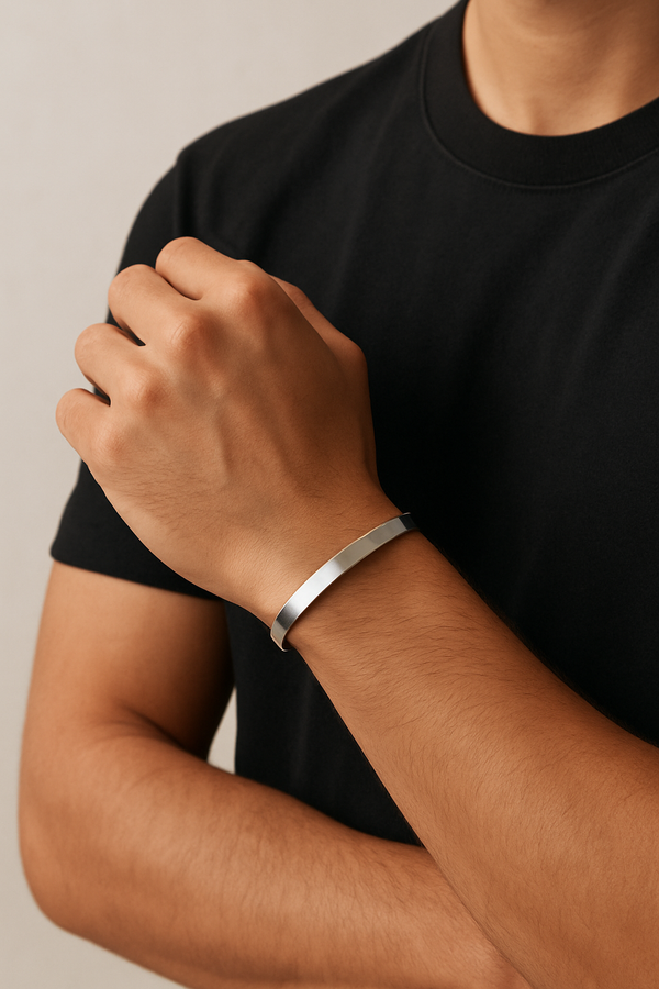 A close-up of a man’s wrist wearing a minimalist silver bracelet, with his hand slightly raised near his chest. He is dressed in a plain black t-shirt, and the bracelet is the focal point against the neutral beige background.