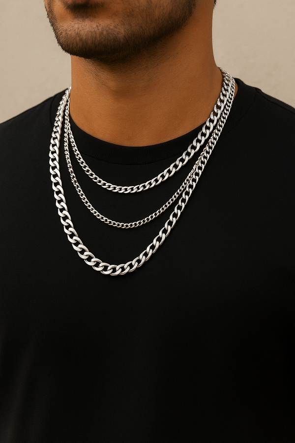 Close-up of a man wearing multiple silver neck chains of different styles and thicknesses over a black t-shirt, showcasing layered jewelry against a neutral background.