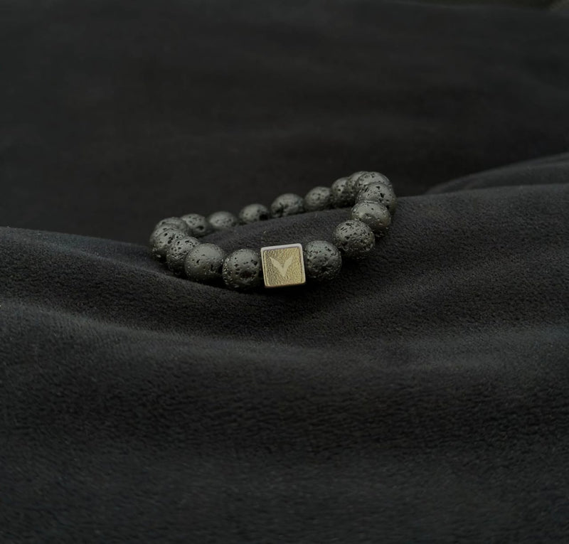 Bracelet with dark beads and a gold square charm on a black fabric background