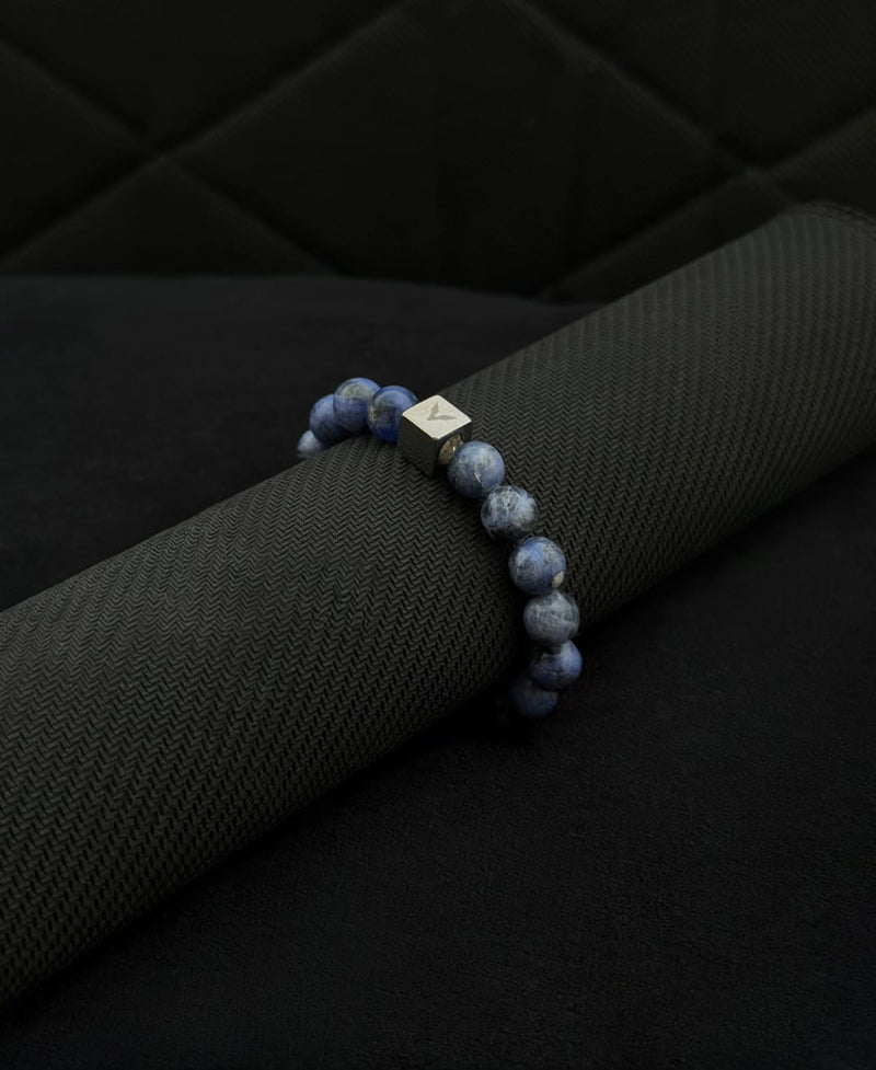 Blue beaded bracelet with a silver cube charm on a black textured surface