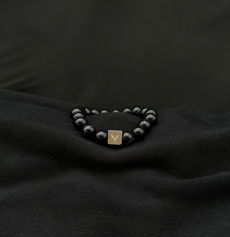Black beaded bracelet with a gold square charm on a dark fabric background