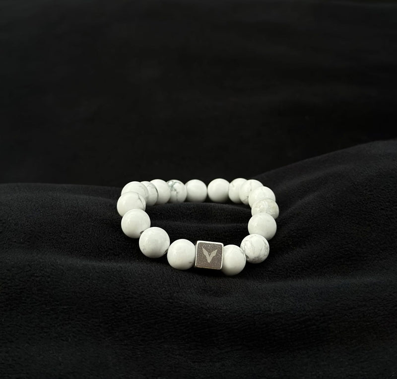 White beaded bracelet with a silver cube charm on a black fabric background
